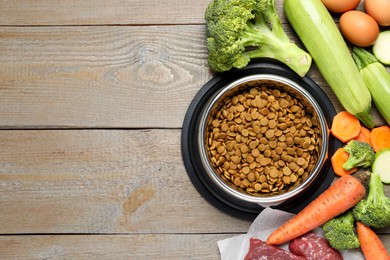 Photo of Dry pet food in feeding bowl and natural products on wooden table, flat lay. Space for text