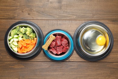 Photo of Natural pet food and chew bone in feeding bowls on wooden table, flat lay