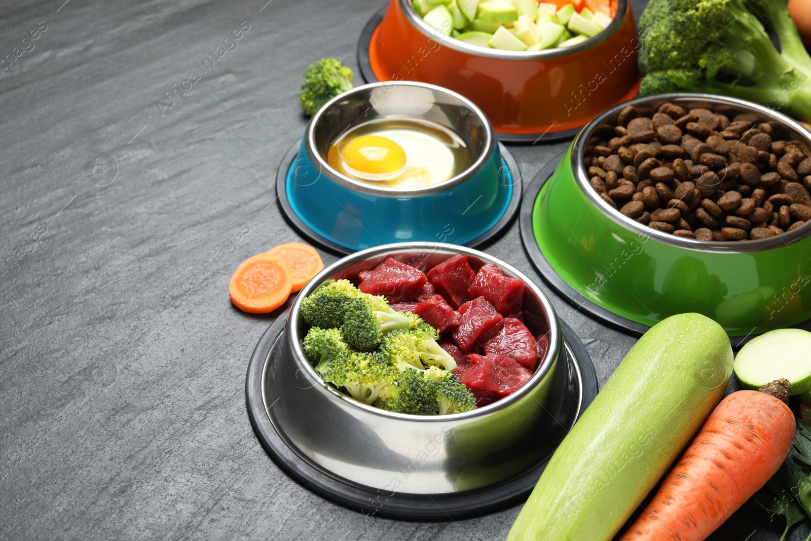 Photo of Natural and dry pet food in feeding bowls among products on dark textured table, closeup. Space for text