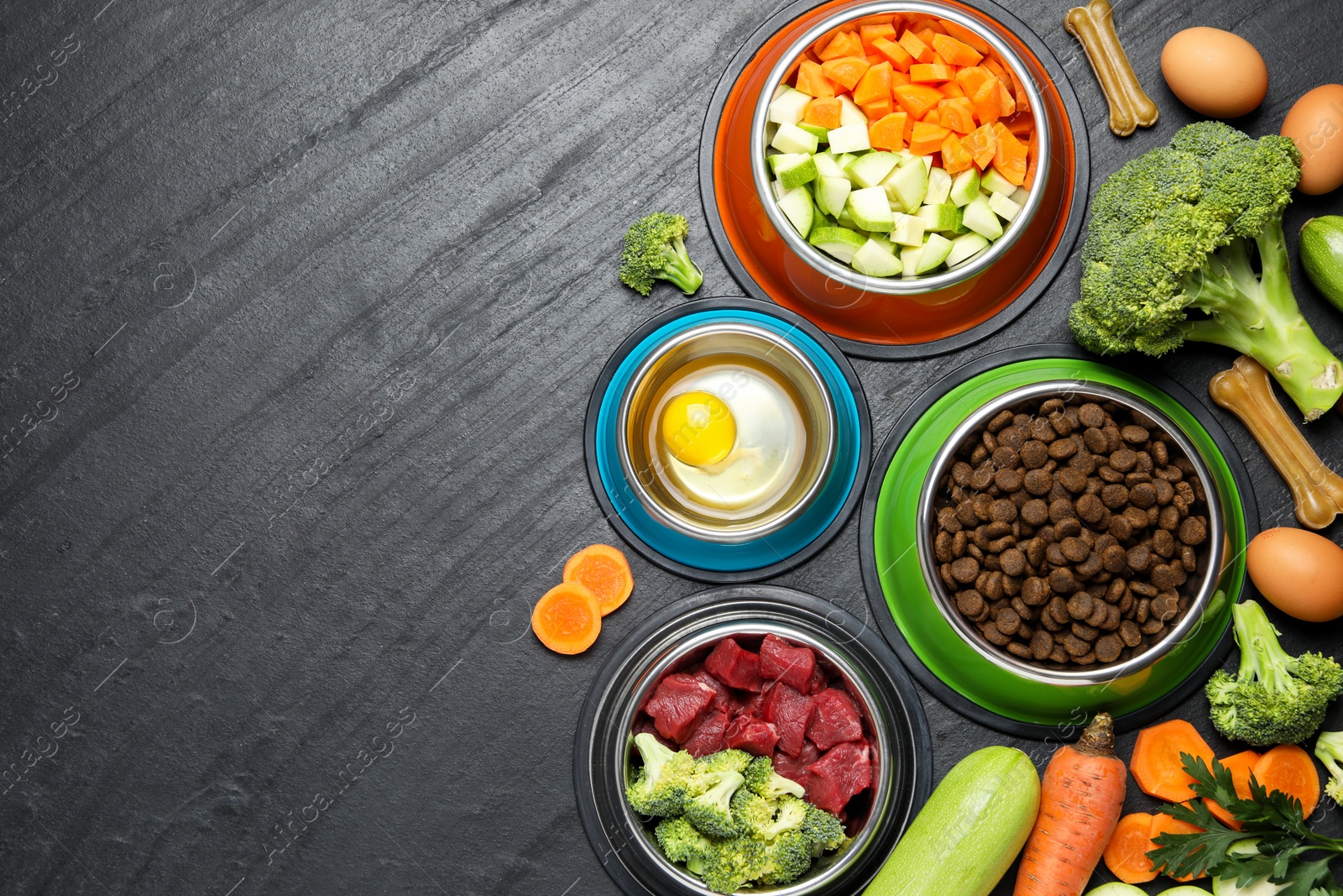 Photo of Natural and dry pet food in feeding bowls among products on dark textured table, flat lay. Space for text