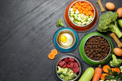 Photo of Natural and dry pet food in feeding bowls among products on dark textured table, flat lay. Space for text