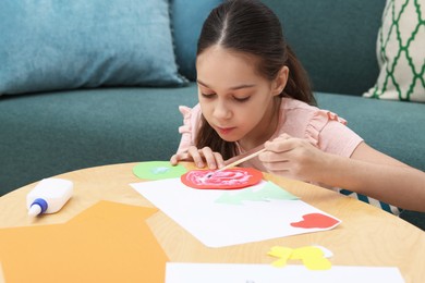 Photo of Girl applying glue onto paper figure for her creative project at table indoors. Art and craft