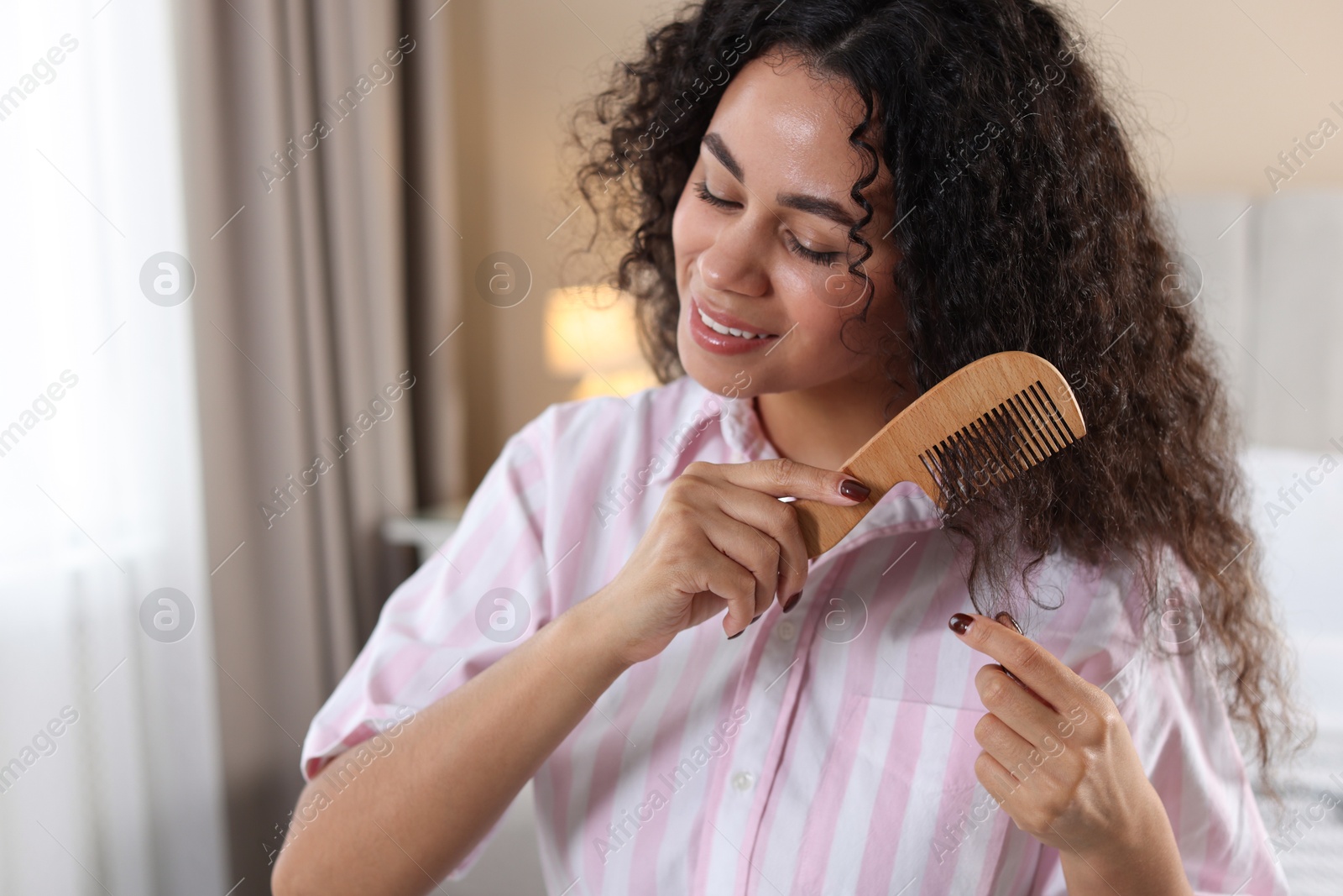 Photo of Smiling young woman brushing her curly hair with comb at home