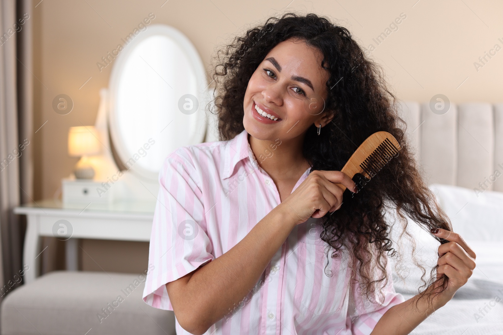 Photo of Smiling young woman brushing her curly hair with comb at home