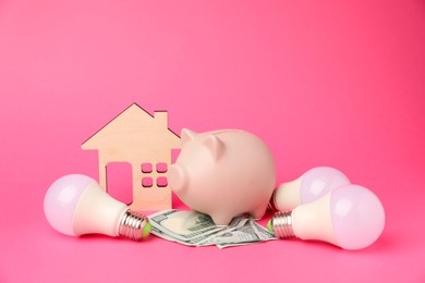 Photo of Piggy bank, dollar banknotes, light bulbs and house model on pink background. Energy saving concept