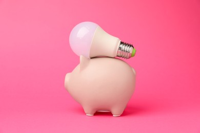 Photo of Piggy bank with light bulb on pink background. Energy saving concept