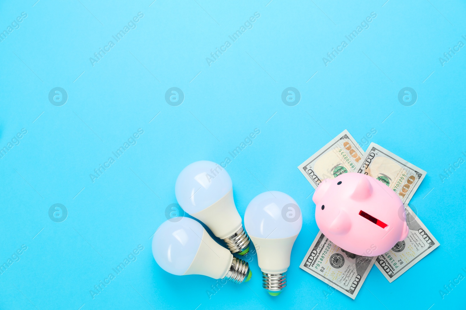 Photo of Piggy bank, dollar banknotes and lightbulbs on light blue background, flat lay with space for text. Energy saving concept