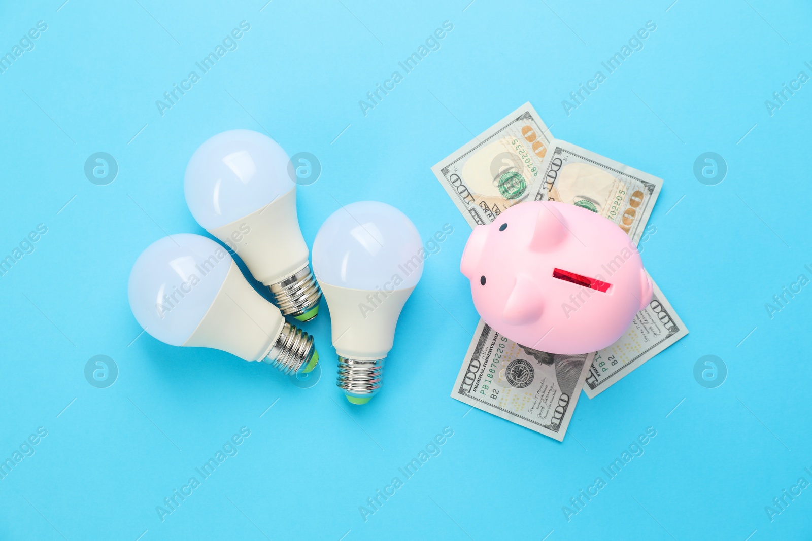 Photo of Piggy bank, dollar banknotes and lightbulbs on light blue background, flat lay. Energy saving concept