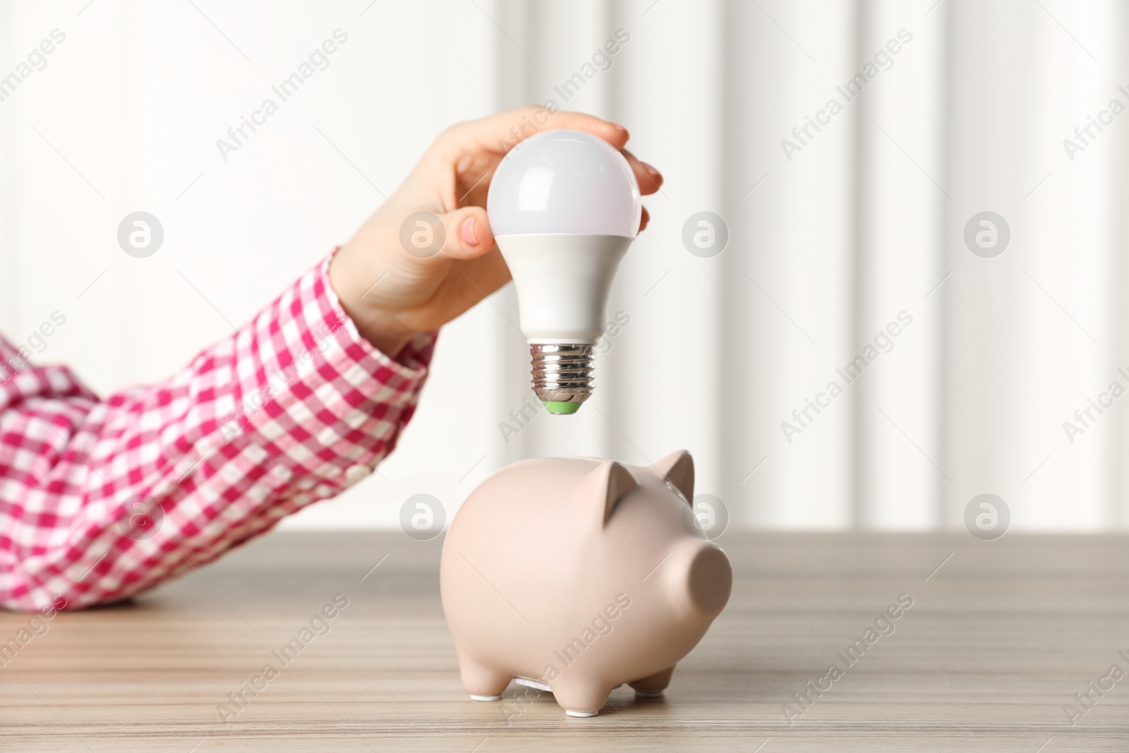 Photo of Woman holding light bulb above piggy bank at wooden table, closeup. Energy saving concept