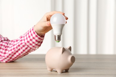 Photo of Woman holding light bulb above piggy bank at wooden table, closeup. Energy saving concept