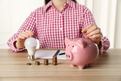 Photo of Woman with light bulb putting coin into piggy bank at wooden table, closeup. Energy saving concept