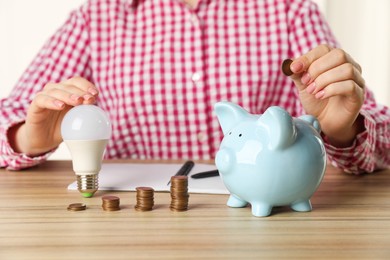 Photo of Woman with light bulb putting coin into piggy bank at wooden table, closeup. Energy saving concept