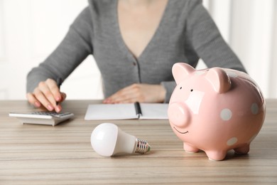 Photo of Woman using calculator at wooden table, focus on piggy bank and light bulb. Energy saving concept