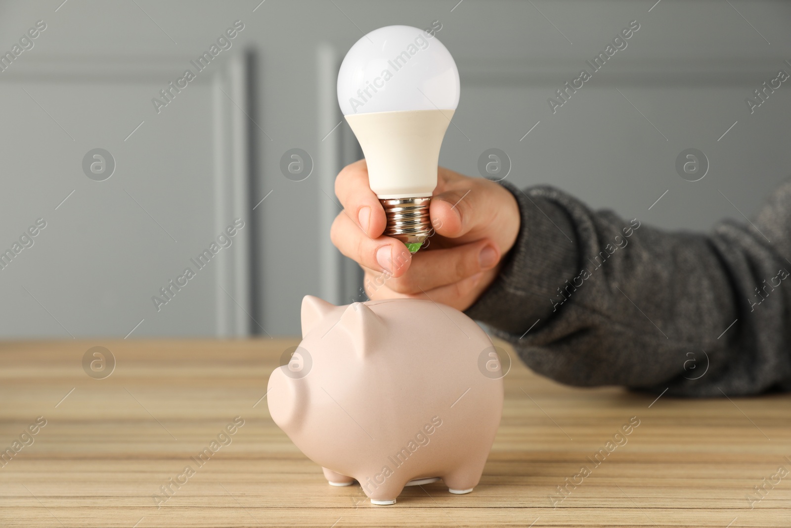 Photo of Man holding light bulb above piggy bank at wooden table, closeup. Energy saving concept