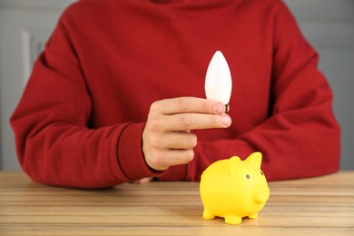 Photo of Man holding light bulb above piggy bank at wooden table, closeup. Energy saving concept