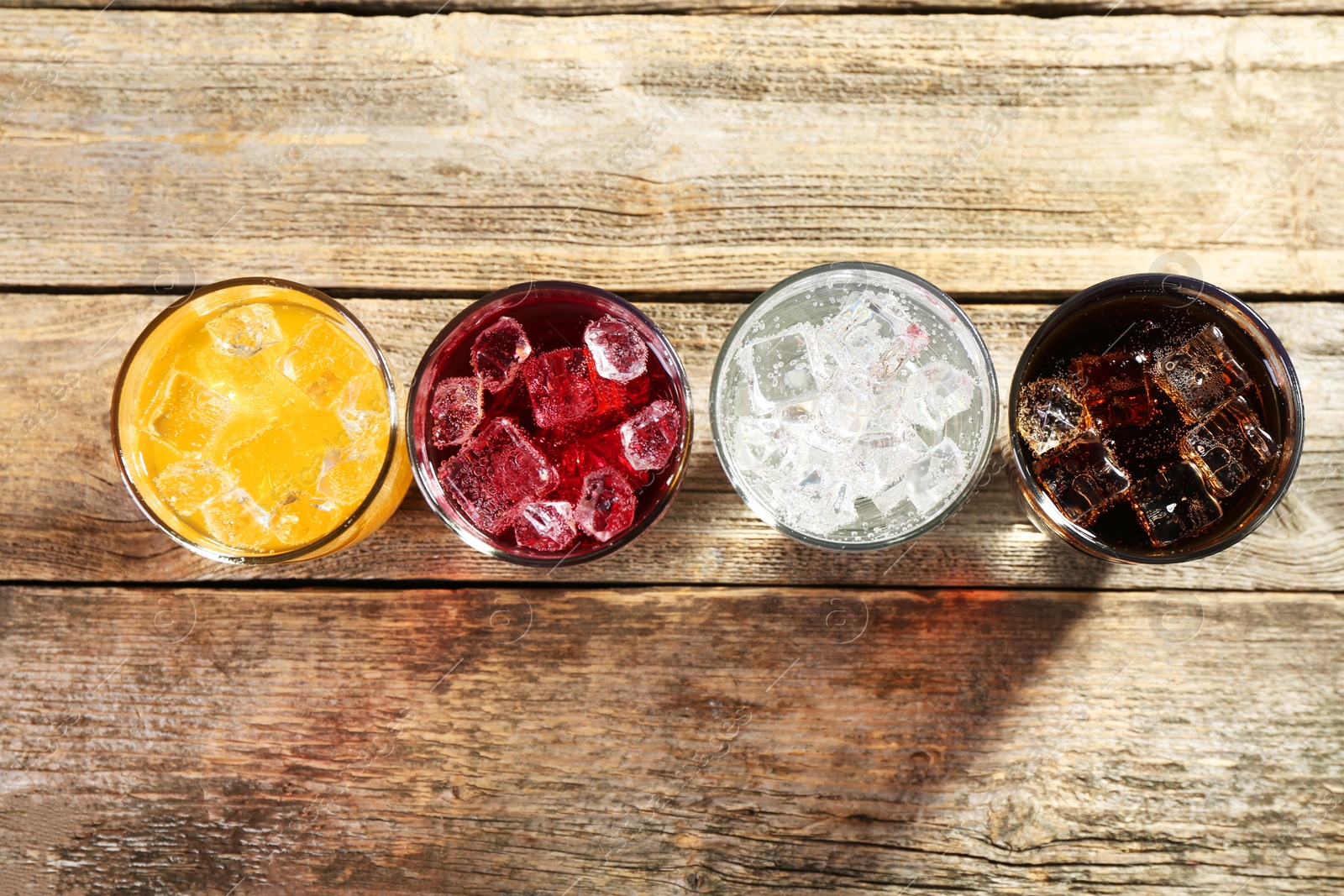 Photo of Soda water of different flavors with ice cubes in glasses on wooden table, flat lay