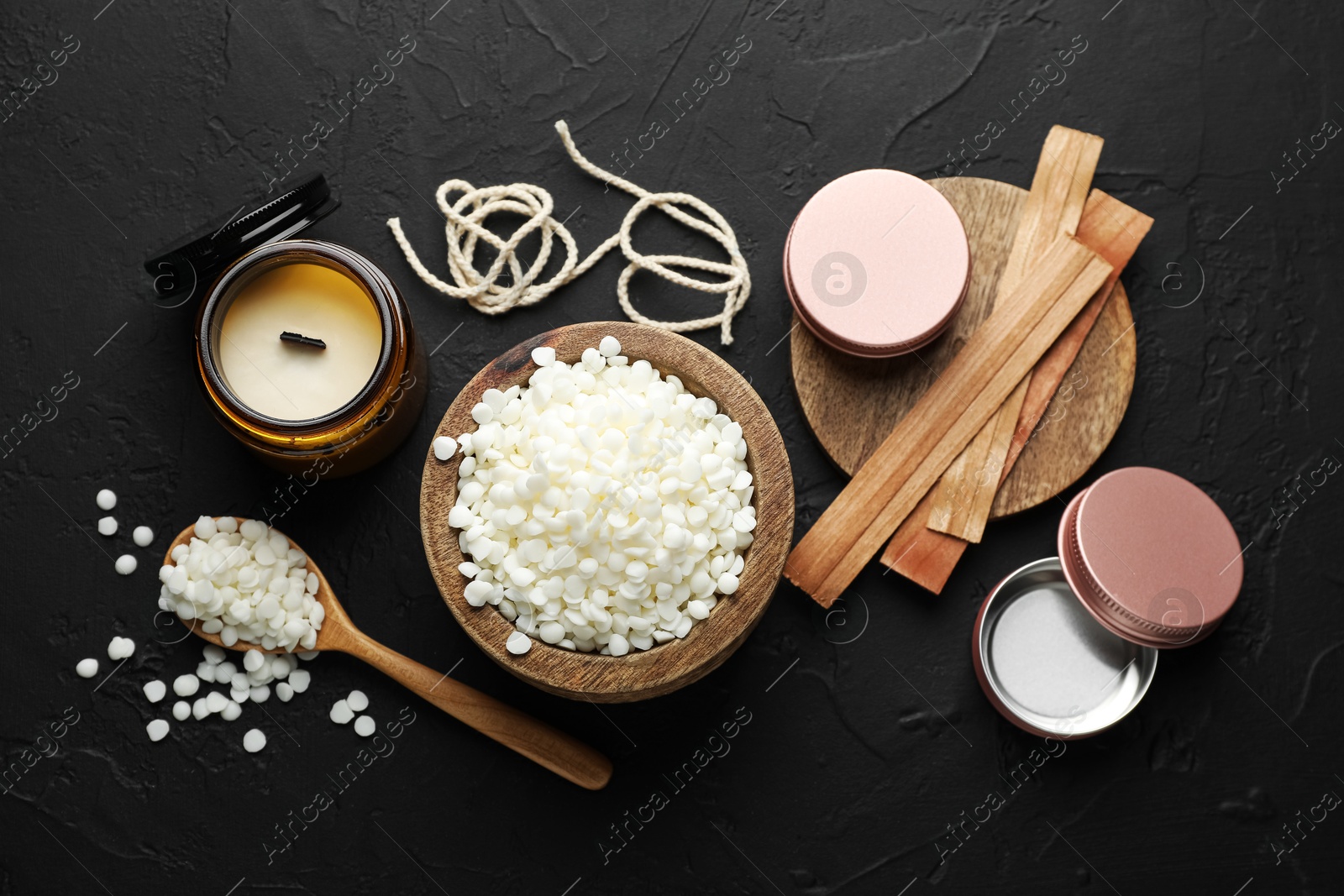 Photo of Soy wax in bowl, candle, wooden wicks, twine and jars on black table, flat lay