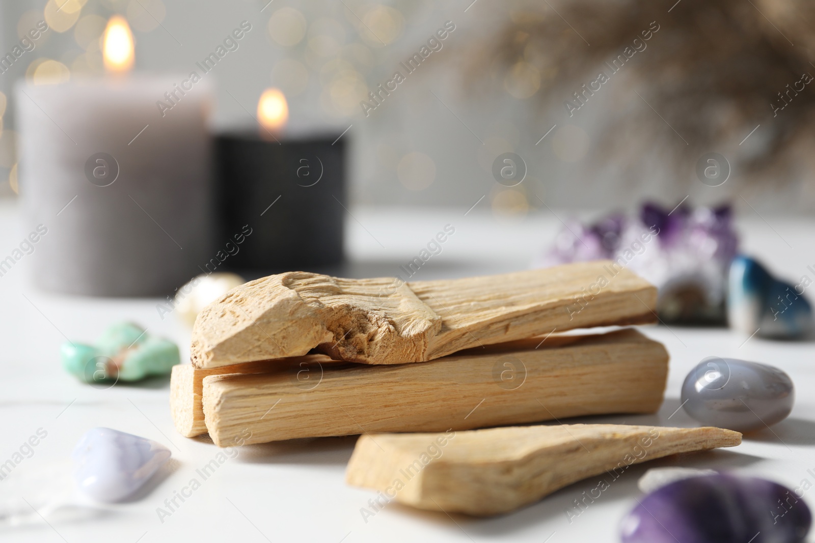 Photo of Palo santo sticks, gemstones and burning candles on white table against blurred lights, closeup