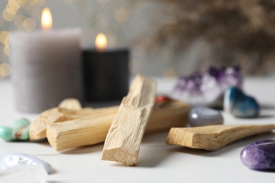 Photo of Palo santo sticks, gemstones and burning candles on white table against blurred lights, closeup