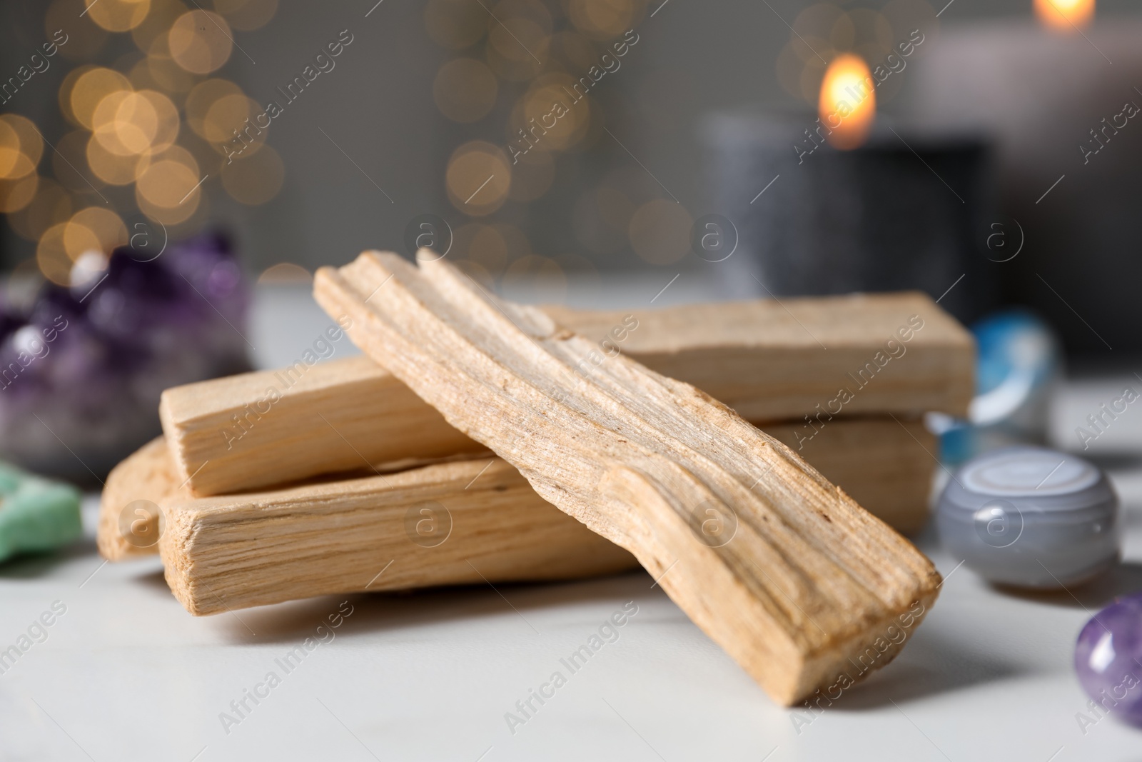 Photo of Palo santo sticks, gemstones and burning candles on white table against blurred lights, closeup