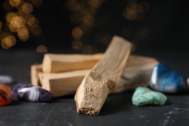 Photo of Palo santo sticks and gemstones on black table against blurred lights, closeup