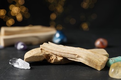 Photo of Palo santo sticks and gemstones on black table against blurred lights, closeup