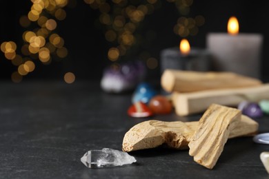 Photo of Palo santo sticks, gemstones and burning candles on black table against blurred lights, closeup. Space for text