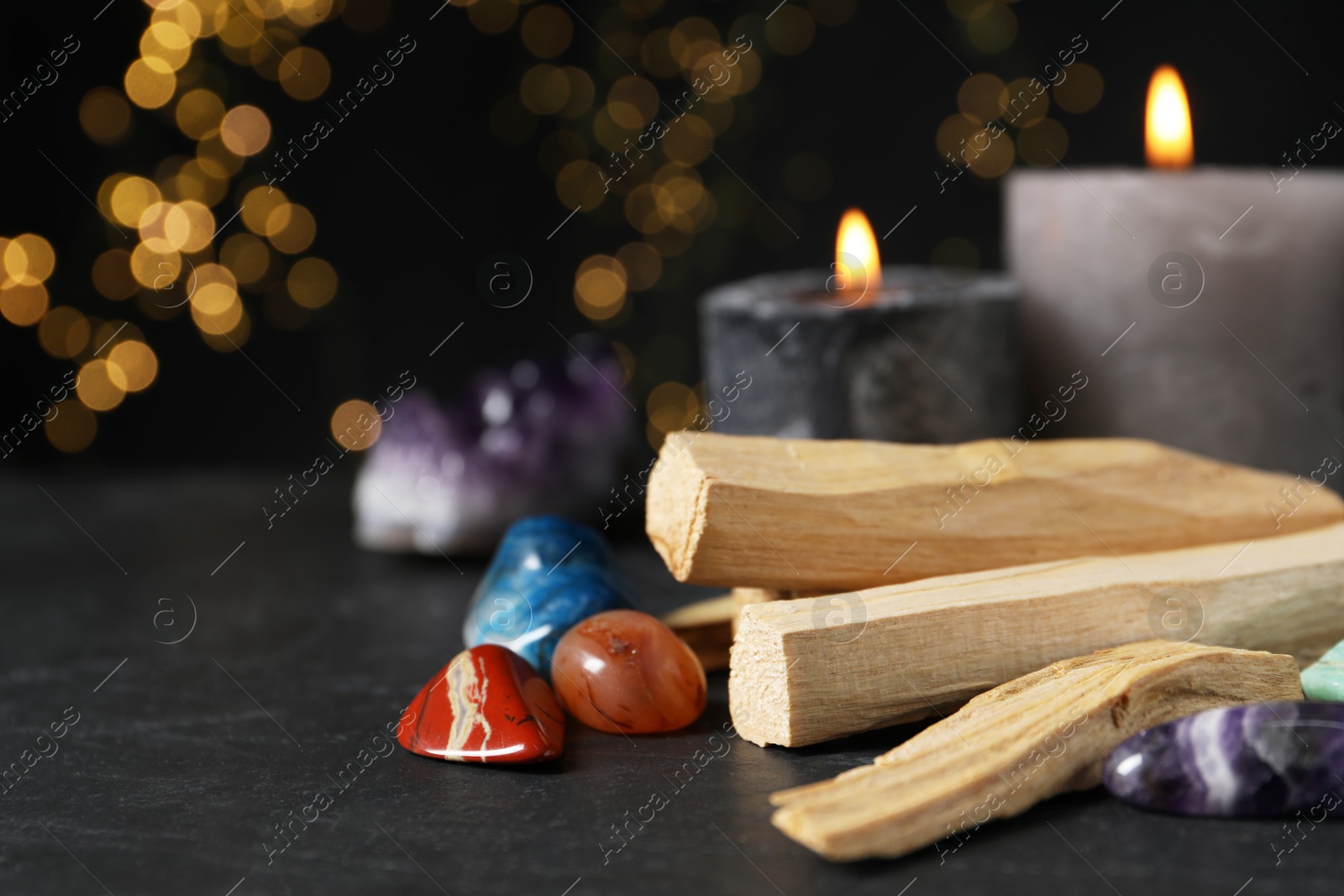 Photo of Palo santo sticks, gemstones and burning candles on black table against blurred lights, closeup. Space for text