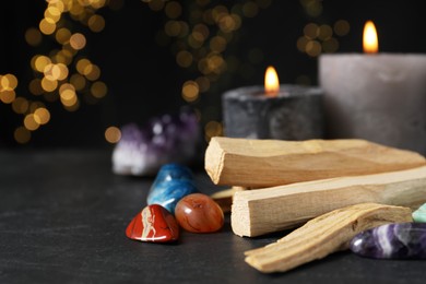 Photo of Palo santo sticks, gemstones and burning candles on black table against blurred lights, closeup. Space for text