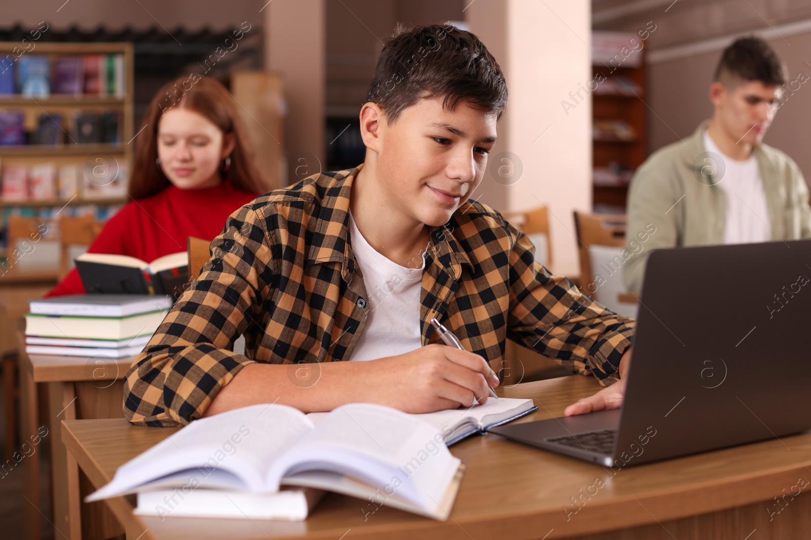 Photo of Boy taking notes while using laptop at desk in public library