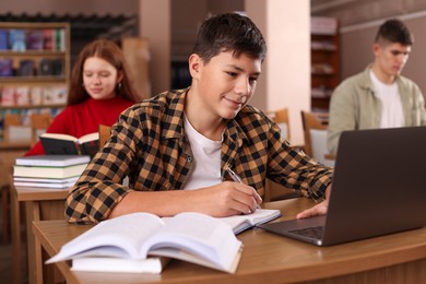 Photo of Boy taking notes while using laptop at desk in public library