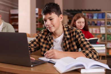 Photo of Boy taking notes while using laptop at desk in public library