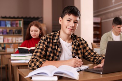 Photo of Boy taking notes while using laptop at desk in public library