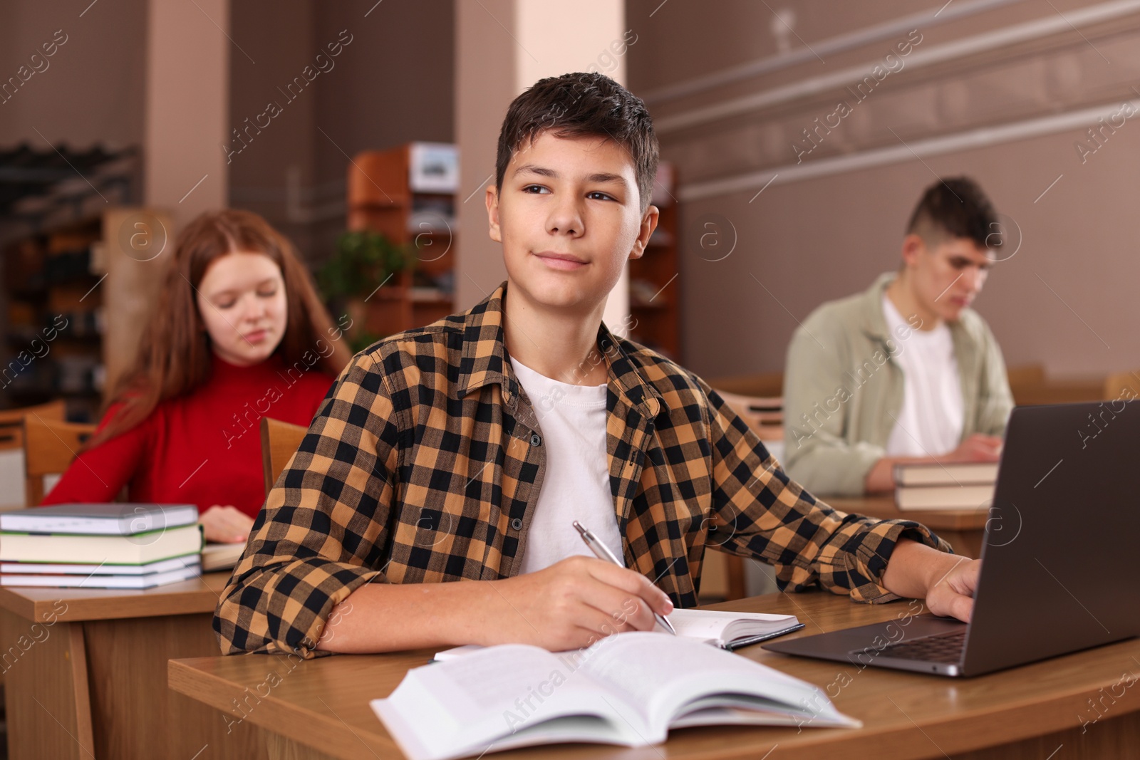 Photo of Boy taking notes while using laptop at desk in public library