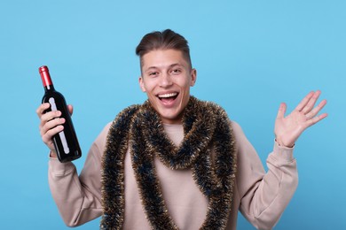 Photo of Happy young man with tinsel and bottle of wine on light blue background