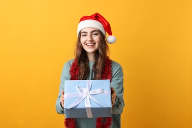 Happy young woman with tinsel, Santa hat and gift box on orange background