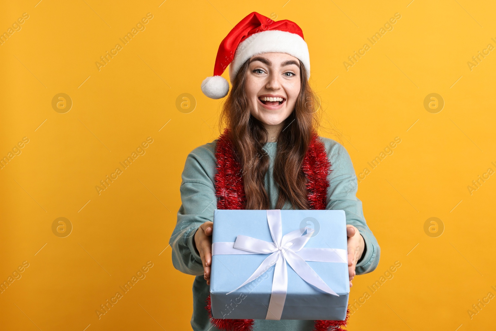 Photo of Happy young woman with tinsel, Santa hat and gift box on orange background