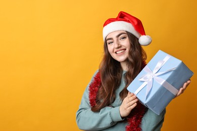 Photo of Happy young woman with tinsel, Santa hat and gift box on orange background. Space for text