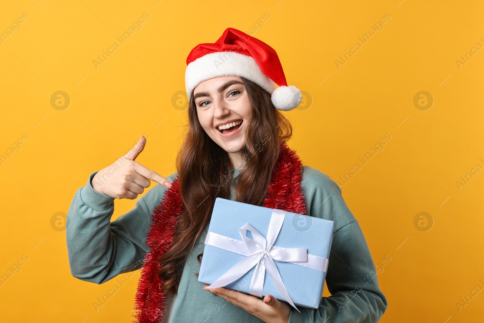 Photo of Happy young woman with tinsel, Santa hat and gift box on orange background