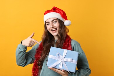 Photo of Happy young woman with tinsel, Santa hat and gift box on orange background