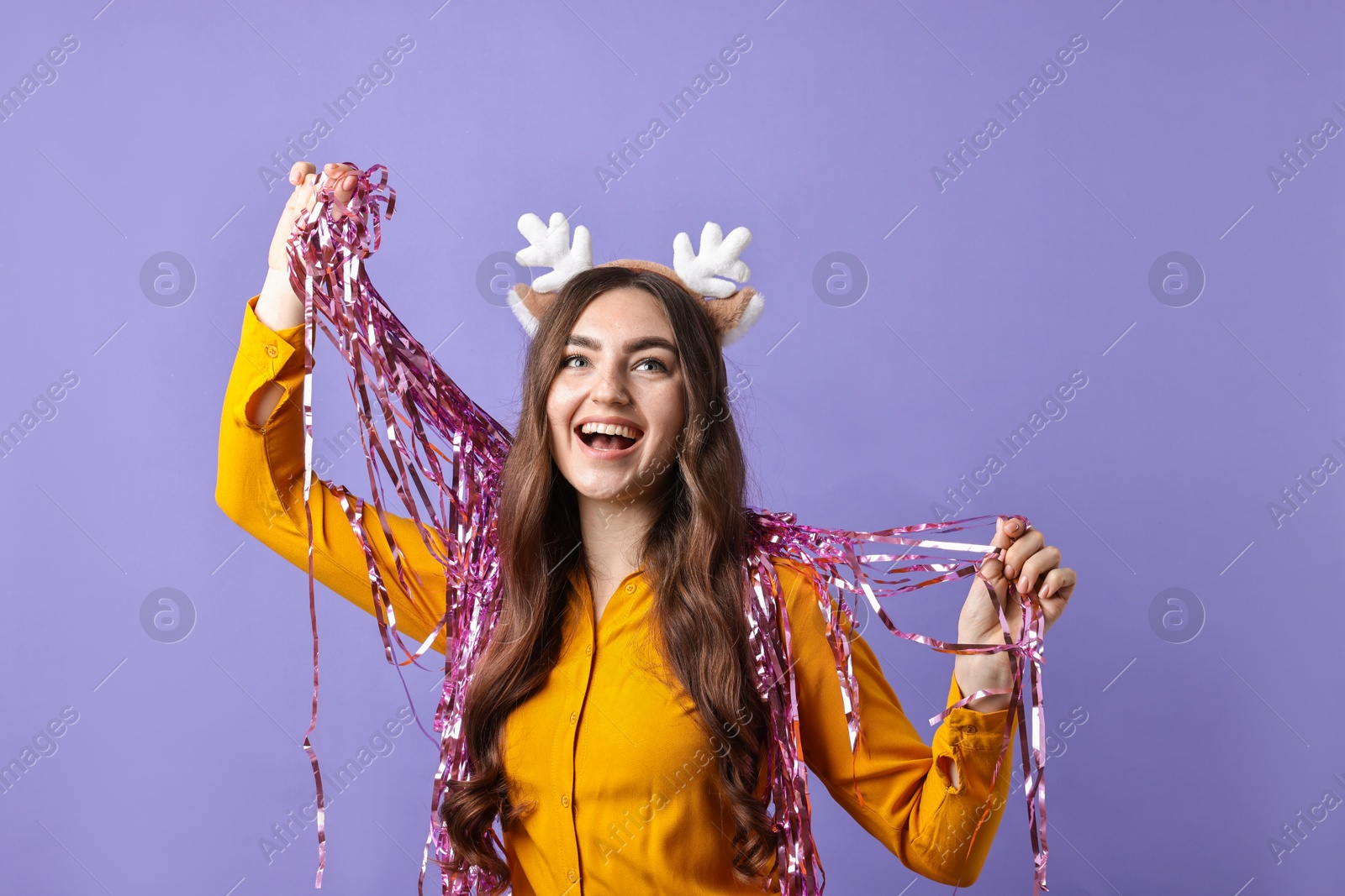 Photo of Happy young woman in reindeer headband with tinsel on purple background