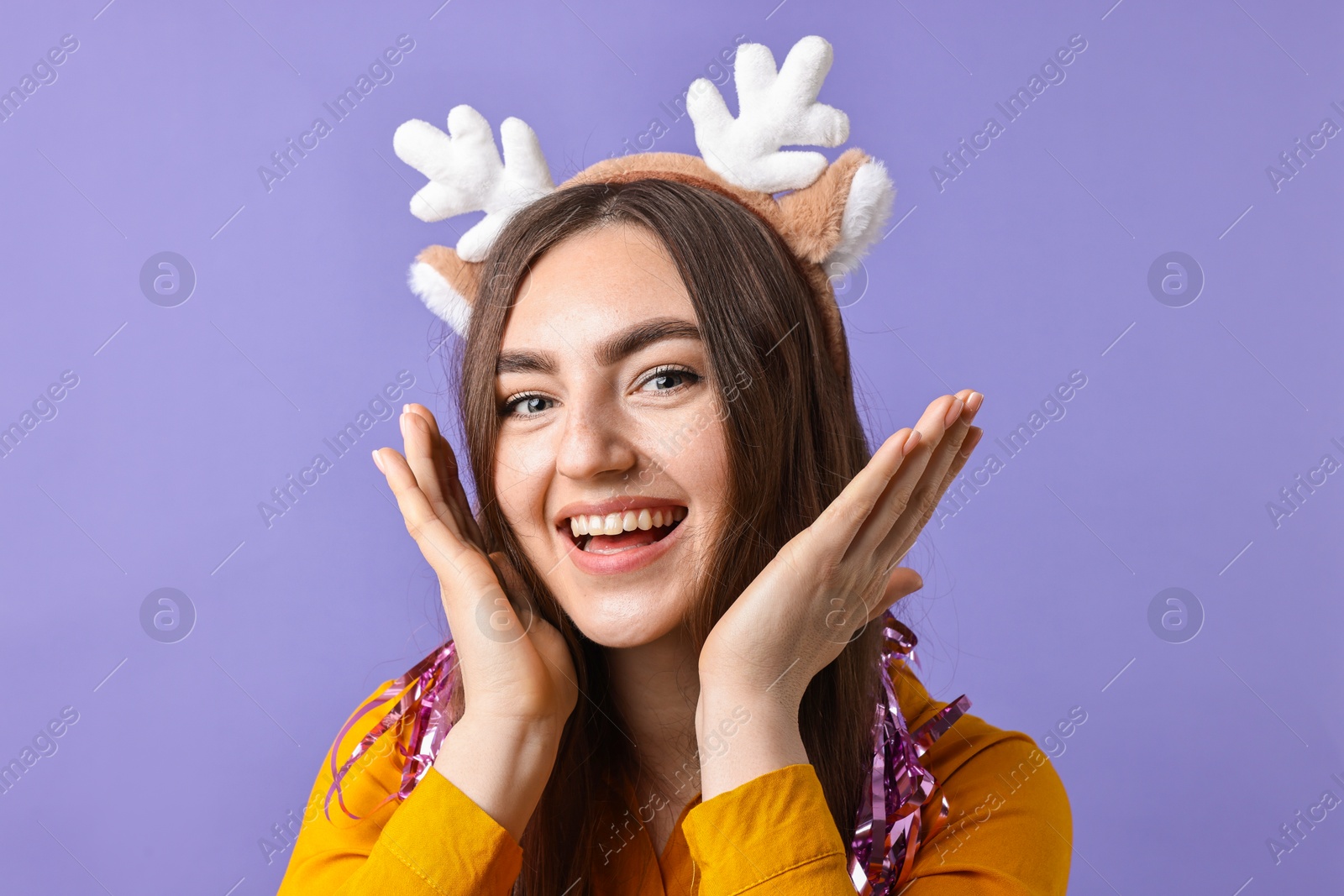 Photo of Happy young woman in reindeer headband with tinsel on purple background