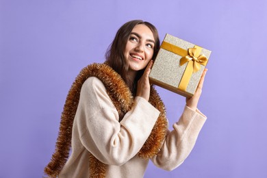 Photo of Happy young woman with tinsel and gift box on purple background