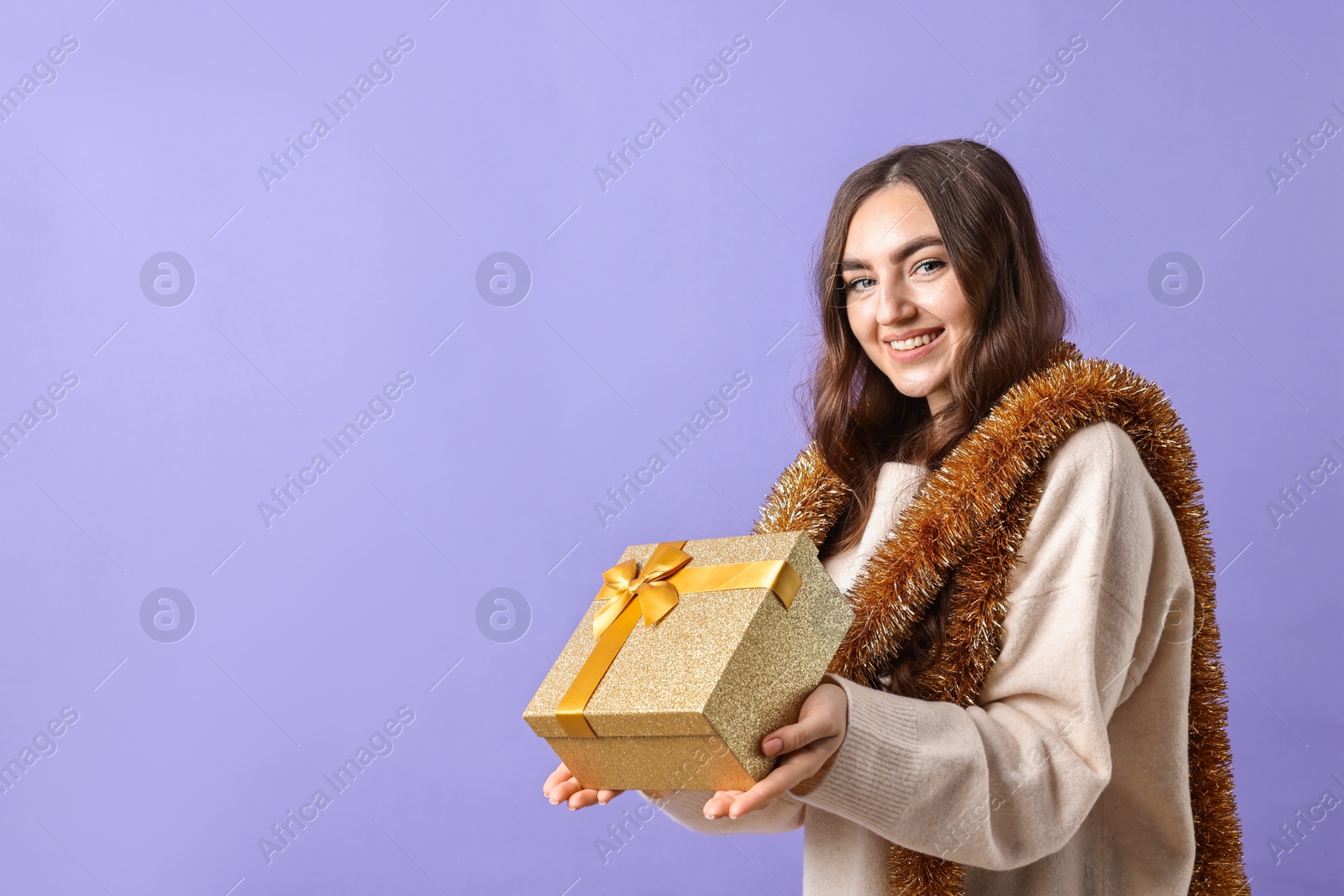 Photo of Happy young woman with tinsel and gift box on purple background. Space for text