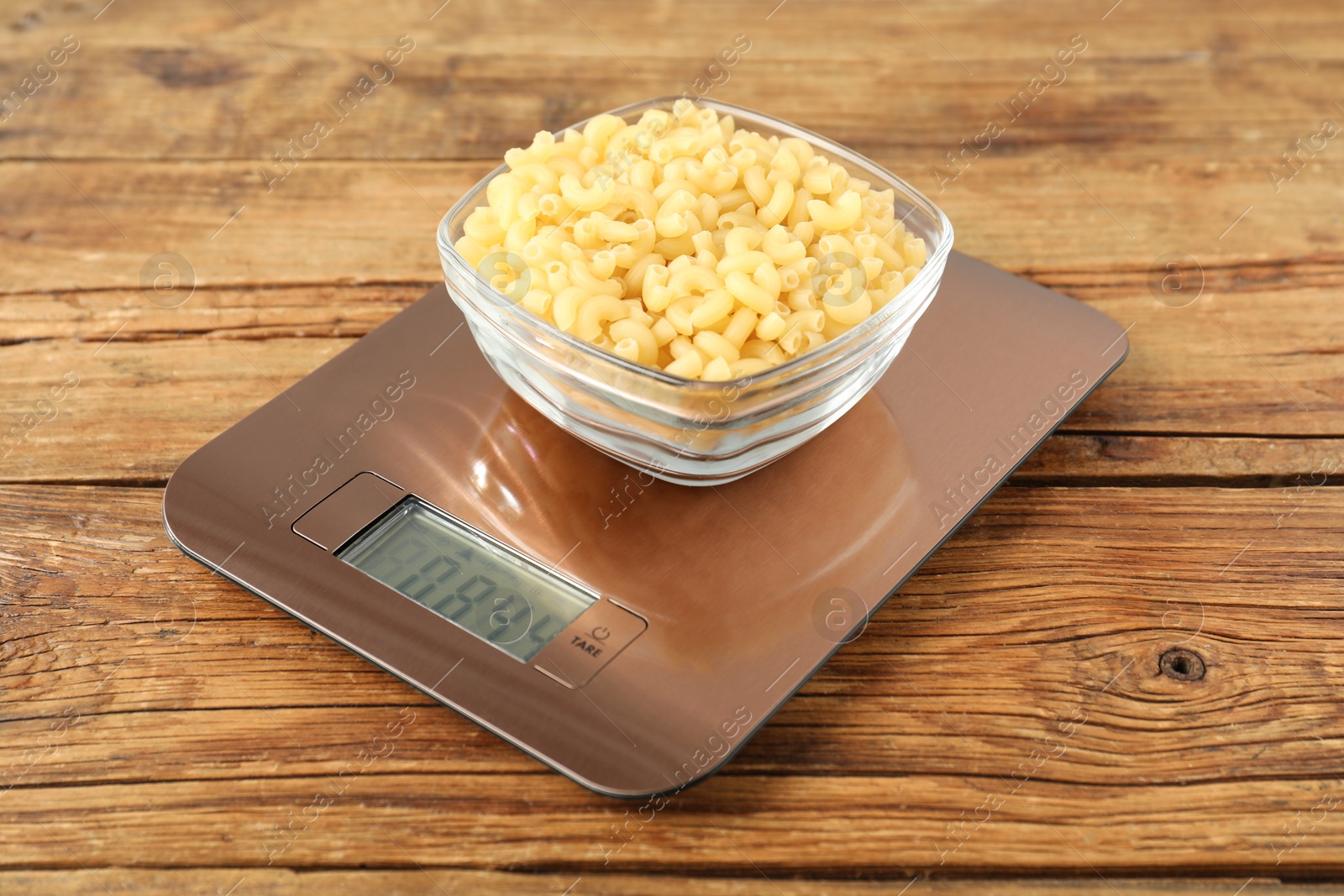 Photo of Modern electronic kitchen scale with bowl of raw pasta on wooden table, closeup