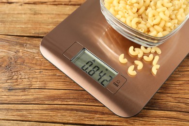 Photo of Modern electronic kitchen scale with bowl of raw pasta on wooden table, closeup