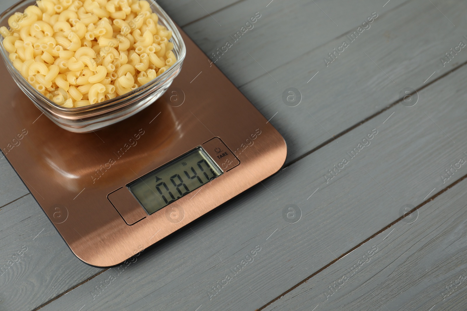 Photo of Modern electronic kitchen scale with bowl of raw pasta on grey wooden table, closeup. Space for text