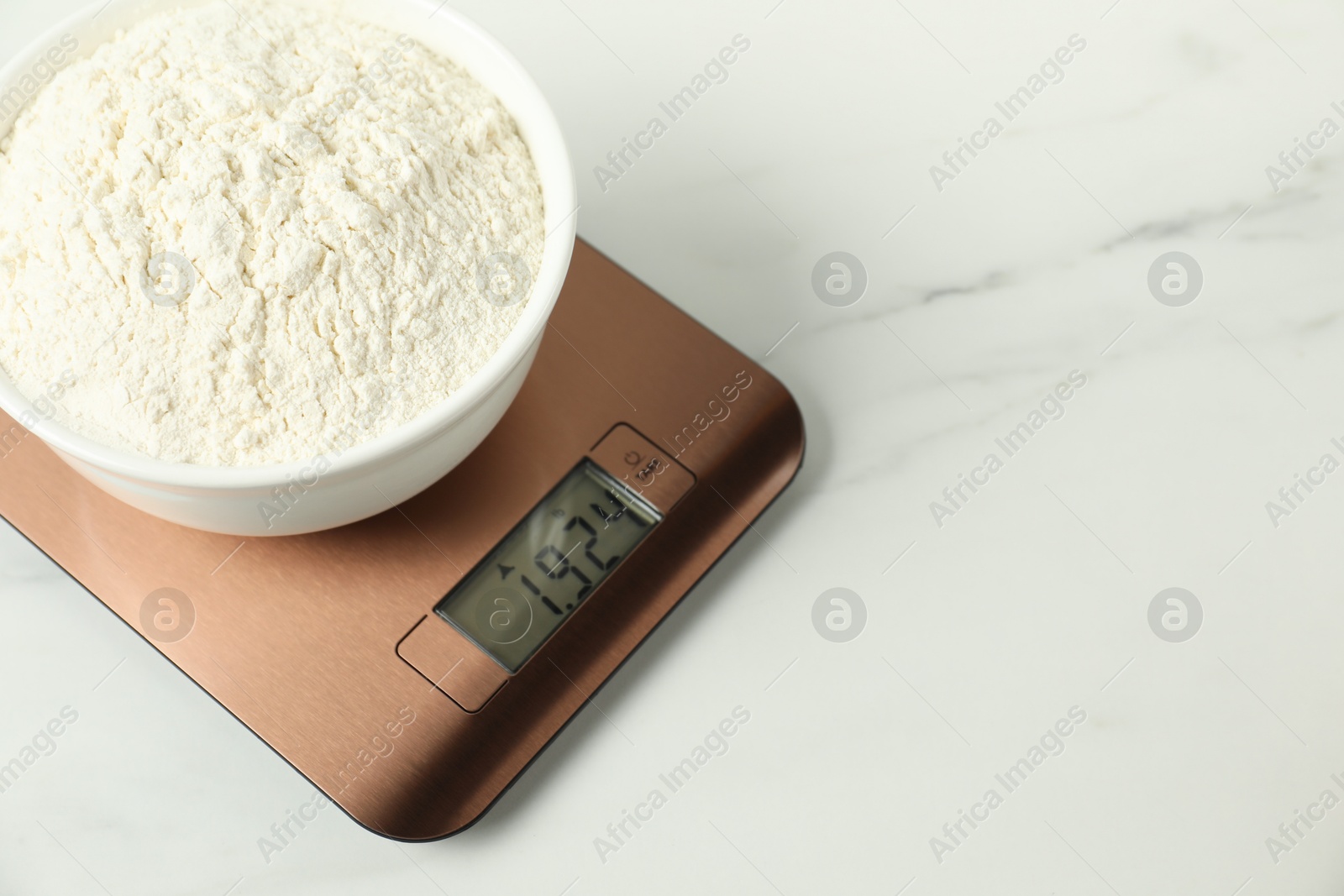 Photo of Modern electronic kitchen scale with bowl of flour on white marble table, closeup. Space for text