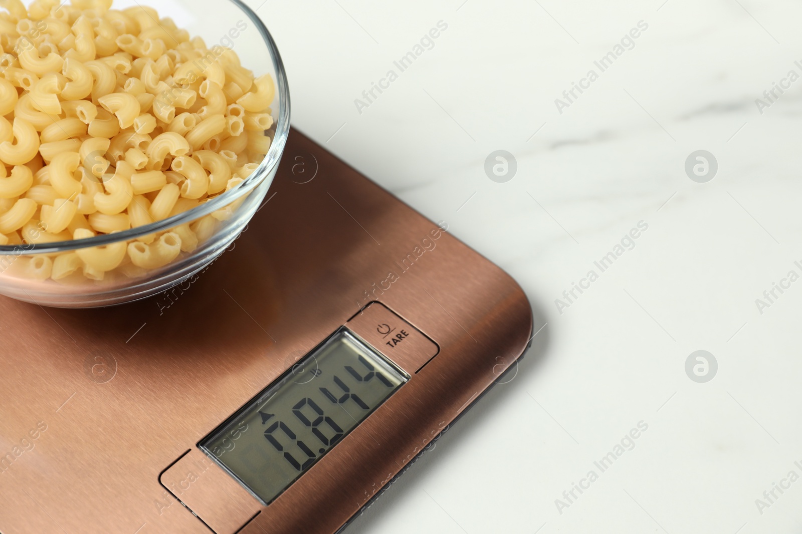 Photo of Modern electronic kitchen scale with bowl of raw pasta on white marble table, closeup. Space for text