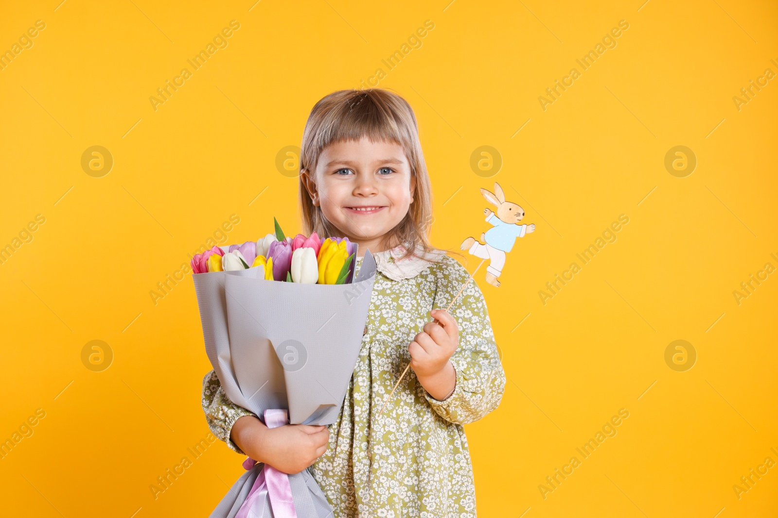 Photo of Smiling little girl with bouquet of tulips and decorative Easter bunny on orange background